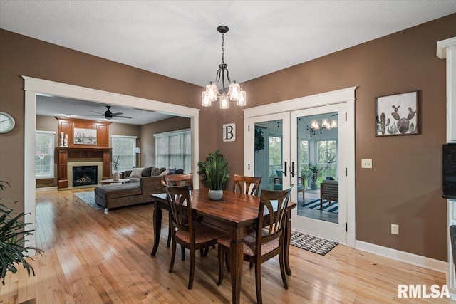 dining area with french doors, baseboards, ceiling fan with notable chandelier, light wood finished floors, and a glass covered fireplace