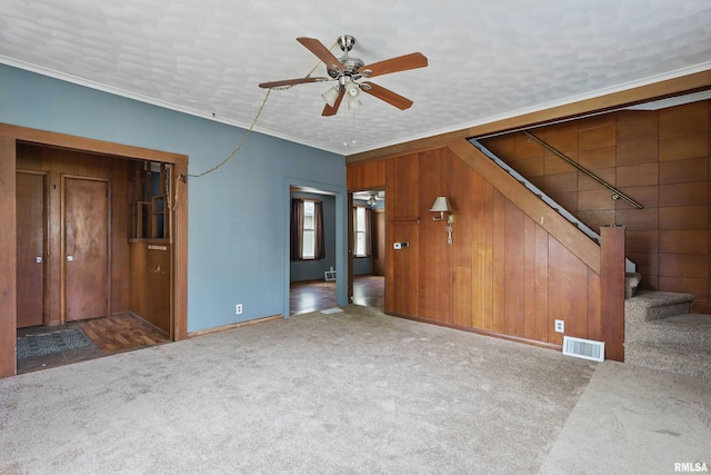 unfurnished living room featuring ceiling fan, wood walls, carpet, visible vents, and ornamental molding
