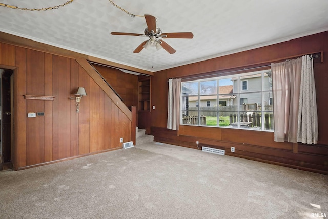 unfurnished living room with ceiling fan, wood walls, carpet, and stairway