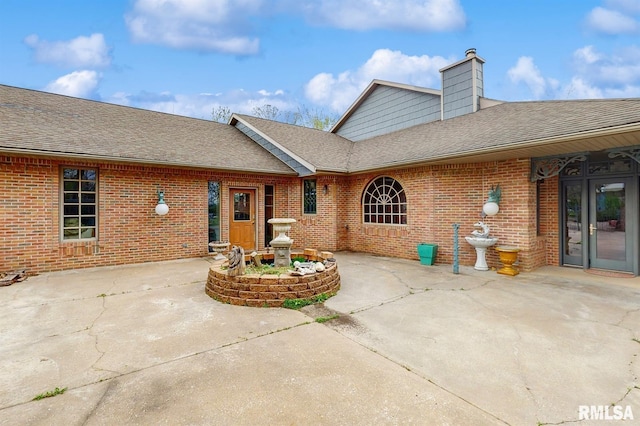 exterior space with brick siding, roof with shingles, a chimney, and a patio
