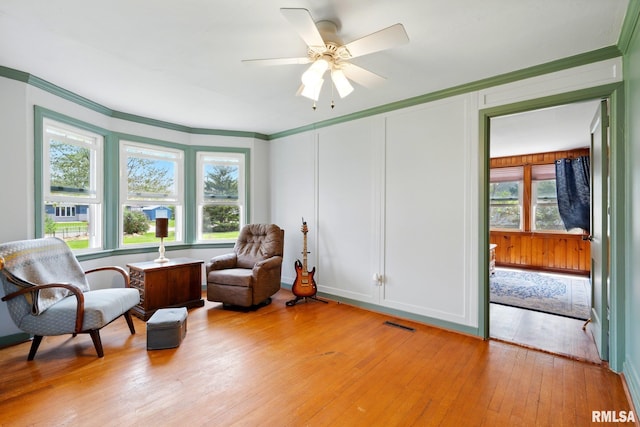 sitting room featuring ceiling fan, visible vents, light wood finished floors, ornamental molding, and a decorative wall