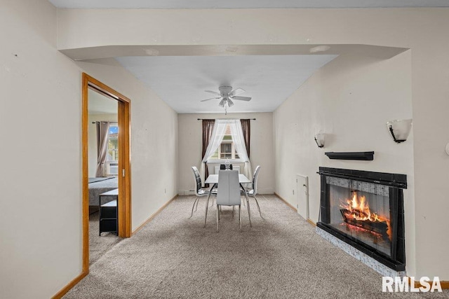 dining space featuring carpet flooring, a warm lit fireplace, baseboards, and ceiling fan