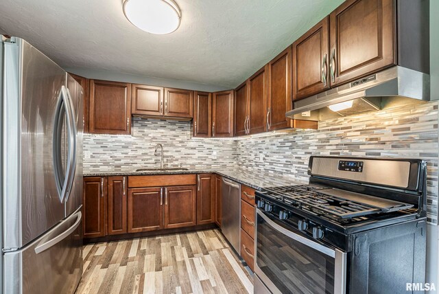 kitchen with appliances with stainless steel finishes, light wood-type flooring, a sink, dark stone counters, and under cabinet range hood