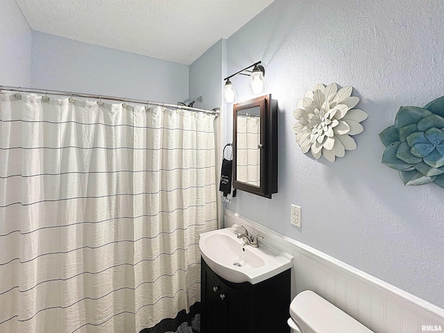 full bathroom featuring a shower with curtain, a textured ceiling, toilet, vanity, and a wainscoted wall