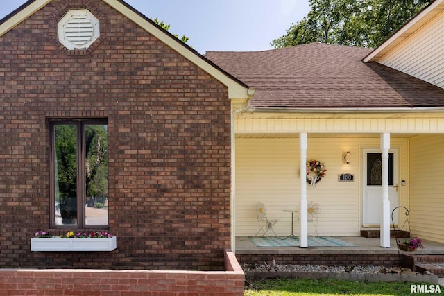 property entrance featuring covered porch, brick siding, and roof with shingles