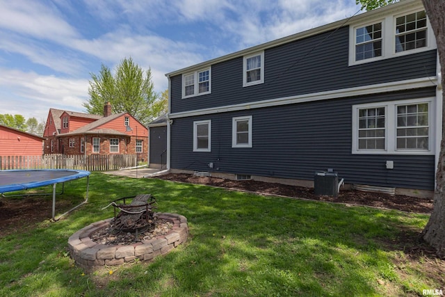 rear view of property featuring a lawn, fence, a trampoline, central air condition unit, and a fire pit