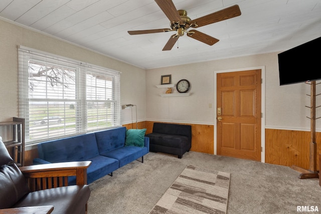 carpeted living room featuring a wainscoted wall, a ceiling fan, wooden walls, and crown molding