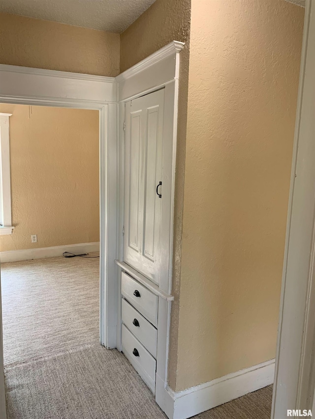 hall featuring light colored carpet, a textured wall, and baseboards