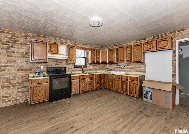 kitchen with black range with electric stovetop, brown cabinetry, light wood finished floors, light countertops, and brick wall