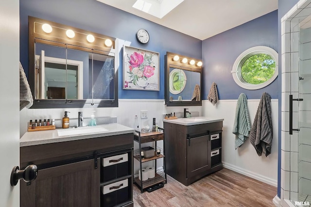 bathroom with a skylight, two vanities, wood finished floors, an enclosed shower, and wainscoting