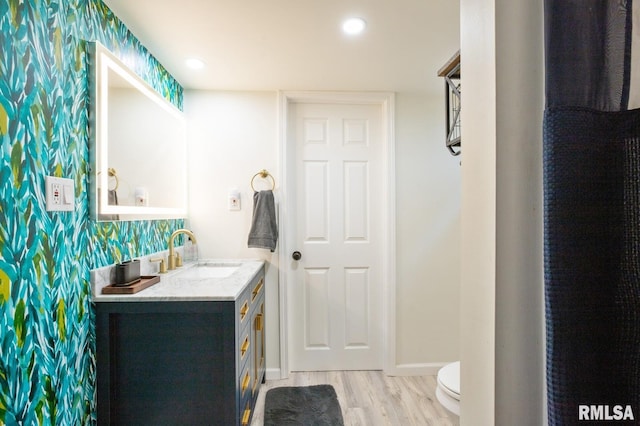 bathroom with wood finished floors, wallpapered walls, toilet, vanity, and baseboards