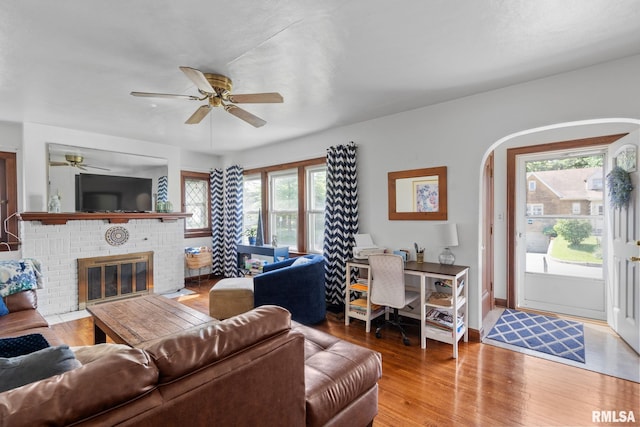 living room featuring a ceiling fan, wood finished floors, a brick fireplace, and arched walkways