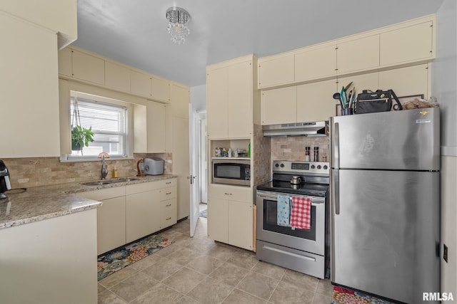 kitchen with appliances with stainless steel finishes, cream cabinets, a sink, under cabinet range hood, and tasteful backsplash