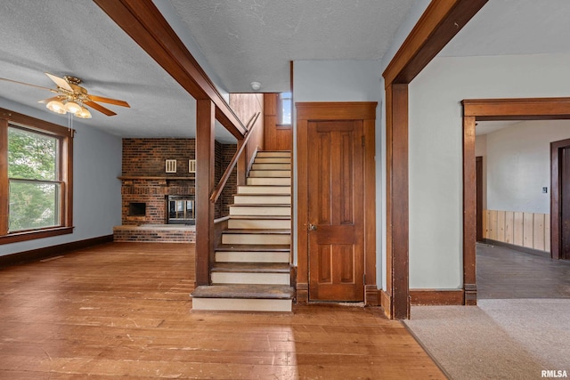 stairs with a textured ceiling, a brick fireplace, hardwood / wood-style flooring, ceiling fan, and baseboards