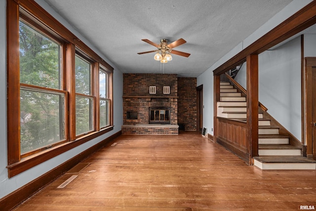 unfurnished living room featuring a textured ceiling, ceiling fan, wood-type flooring, a brick fireplace, and baseboards