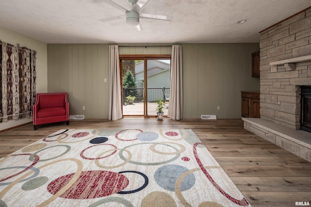 sitting room with wood finished floors, a stone fireplace, a ceiling fan, a textured ceiling, and wood walls