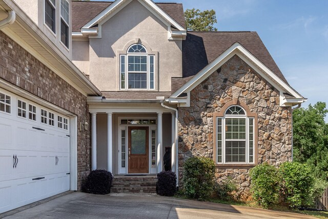 view of exterior entry with a garage, stone siding, stucco siding, a shingled roof, and concrete driveway