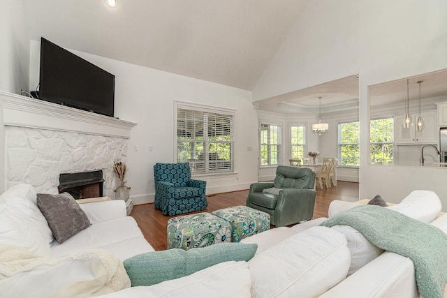 living room with a stone fireplace, wood finished floors, high vaulted ceiling, baseboards, and an inviting chandelier