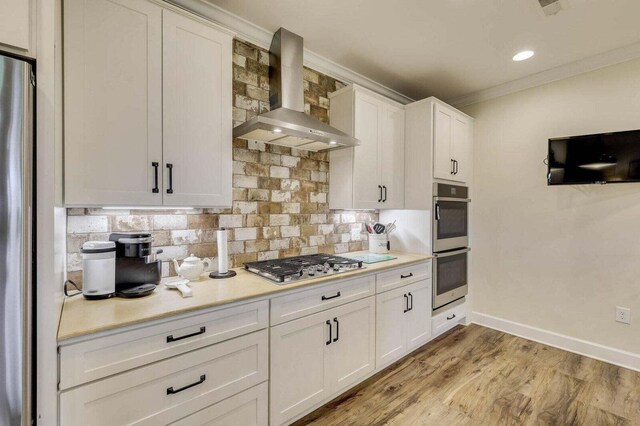 kitchen with appliances with stainless steel finishes, backsplash, wall chimney range hood, light hardwood / wood-style floors, and white cabinetry