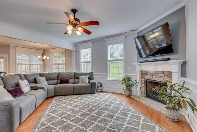 living room featuring light wood-style floors, a chandelier, a ceiling fan, a stone fireplace, and crown molding