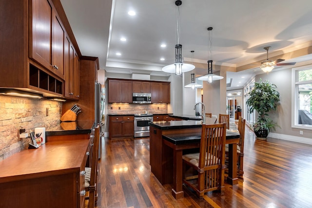 kitchen with tasteful backsplash, stainless steel appliances, ceiling fan, hanging light fixtures, and an island with sink