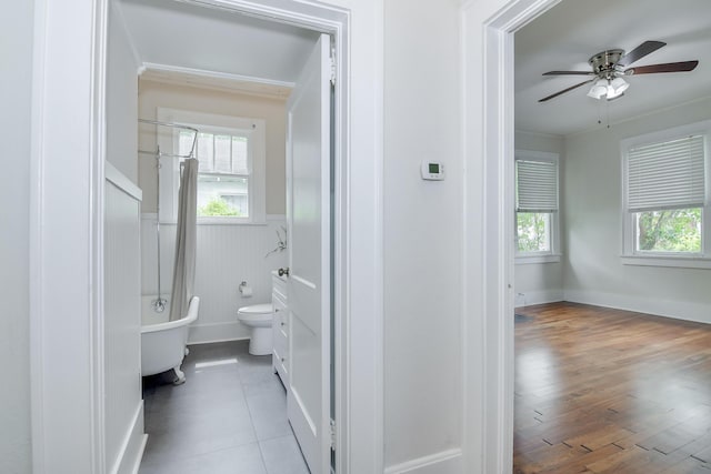 full bath featuring a soaking tub, a wealth of natural light, ceiling fan, and wainscoting