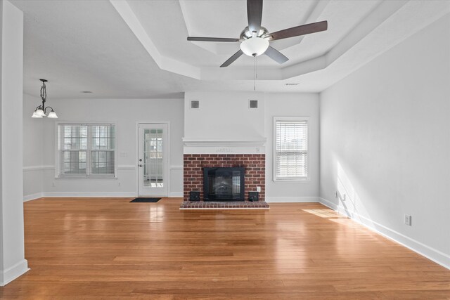 unfurnished living room with a brick fireplace, a tray ceiling, ceiling fan with notable chandelier, and light hardwood / wood-style floors