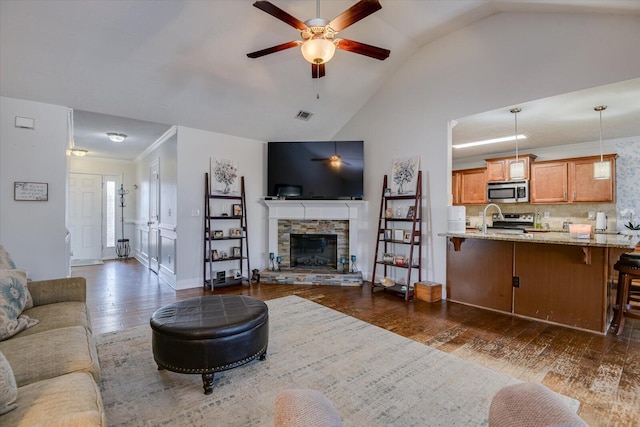 living room with a stone fireplace, ceiling fan, dark wood-type flooring, vaulted ceiling, and sink