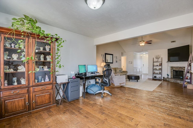 office with a textured ceiling, lofted ceiling, wood-type flooring, a stone fireplace, and ceiling fan