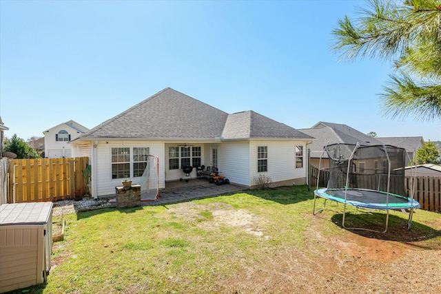 back of property with a trampoline, fence, roof with shingles, a yard, and a patio area