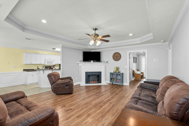 living area with a ceiling fan, light wood finished floors, a tray ceiling, a glass covered fireplace, and crown molding