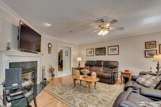 living room with ceiling fan, crown molding, wood-type flooring, a textured ceiling, and a tiled fireplace