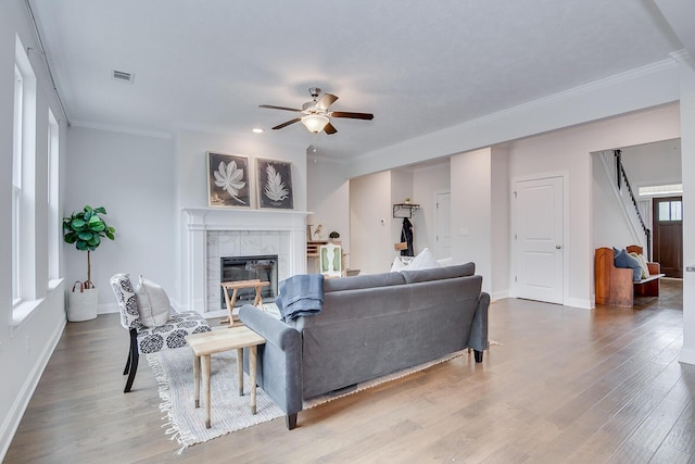 living area with a ceiling fan, a tile fireplace, wood finished floors, crown molding, and stairs