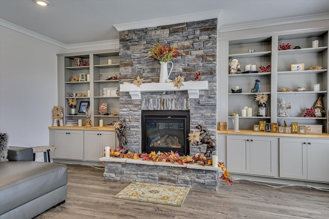 living room with built in shelves, a stone fireplace, wood-type flooring, and ornamental molding