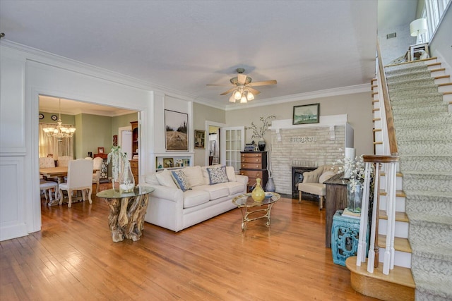 living room featuring hardwood / wood-style flooring, crown molding, ceiling fan with notable chandelier, and a brick fireplace