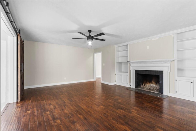 unfurnished living room featuring built in shelves, dark wood-type flooring, ceiling fan, baseboards, and a barn door