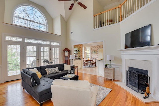 living room featuring ceiling fan, french doors, light hardwood / wood-style flooring, a towering ceiling, and a tiled fireplace