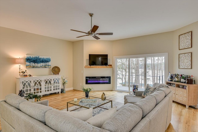 living room with ceiling fan, light wood-type flooring, a glass covered fireplace, and baseboards