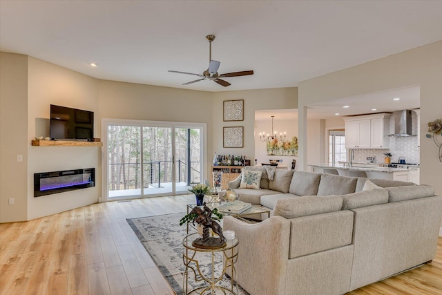 living room featuring ceiling fan with notable chandelier, light wood-type flooring, a glass covered fireplace, and recessed lighting