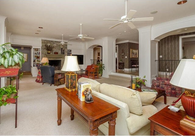 carpeted living room featuring ceiling fan, a stone fireplace, crown molding, and decorative columns