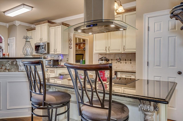 bar with backsplash, dark stone countertops, island range hood, and cream cabinetry