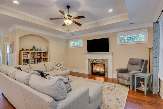 living room with ceiling fan, dark wood-type flooring, a raised ceiling, a stone fireplace, and crown molding