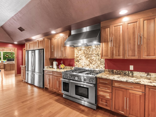 kitchen with appliances with stainless steel finishes, tasteful backsplash, light stone counters, vaulted ceiling, and wall chimney range hood