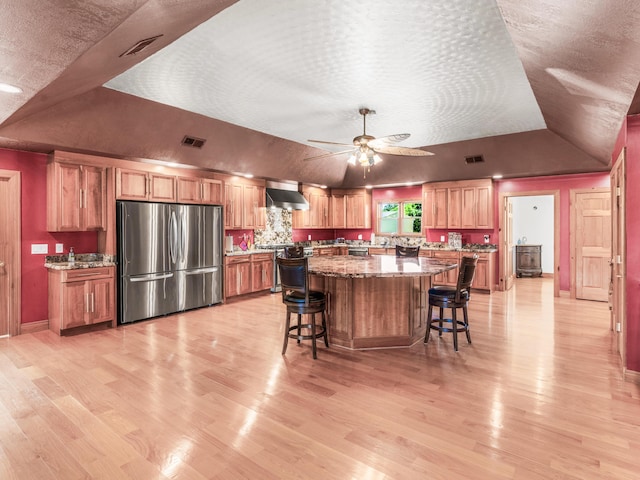 kitchen featuring a center island, wall chimney range hood, ceiling fan, stainless steel fridge, and light stone countertops