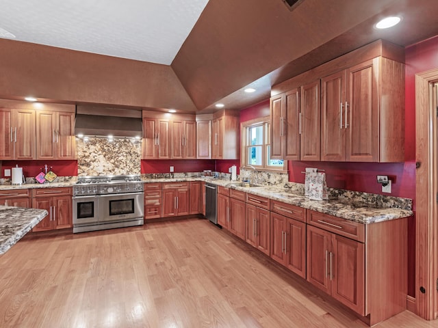 kitchen with sink, vaulted ceiling, wall chimney exhaust hood, light stone counters, and stainless steel appliances