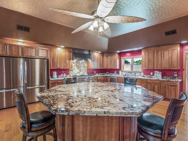 kitchen featuring wall chimney exhaust hood, light hardwood / wood-style floors, appliances with stainless steel finishes, a kitchen island, and light stone counters