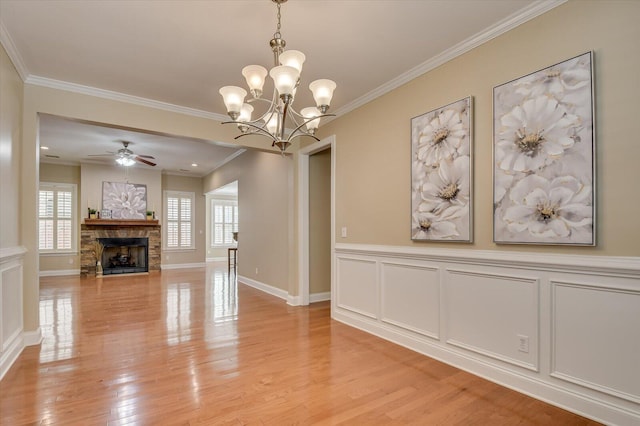 interior space with crown molding, light wood-type flooring, a stone fireplace, ceiling fan with notable chandelier, and a decorative wall