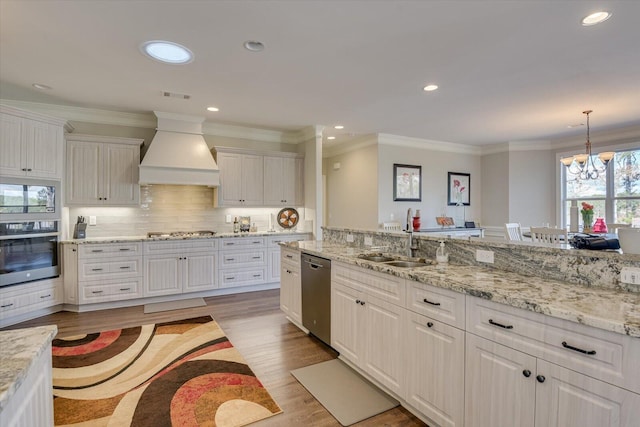 kitchen with stainless steel appliances, a sink, white cabinets, hanging light fixtures, and custom range hood