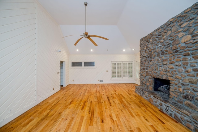 unfurnished living room with vaulted ceiling, ceiling fan, light hardwood / wood-style flooring, a stone fireplace, and wood walls