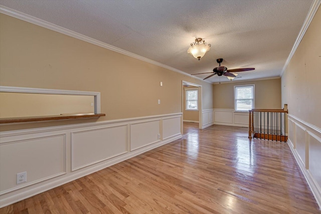 unfurnished room with a textured ceiling, light wood-style flooring, a ceiling fan, wainscoting, and crown molding